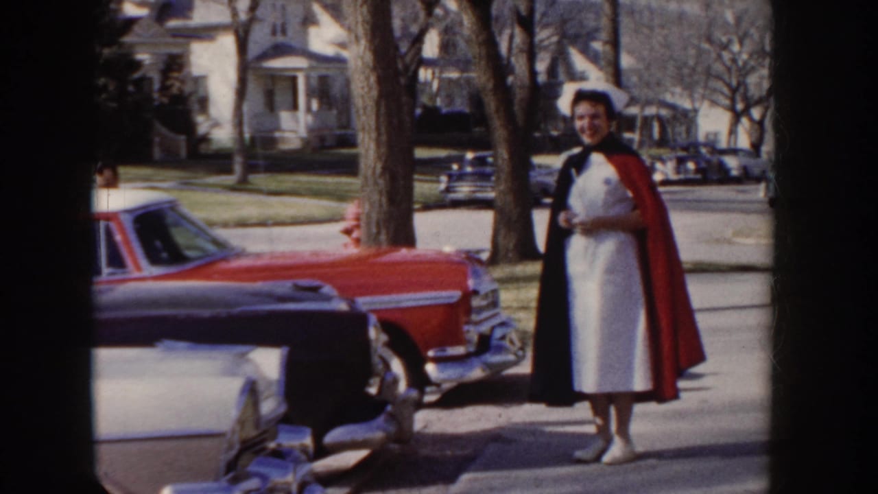 1955 Idaho: nursing graduate in white dress, dark cape, and nurse's cap stands beside a large-finned red American sedan on a tree-lined suburban sidewalk.