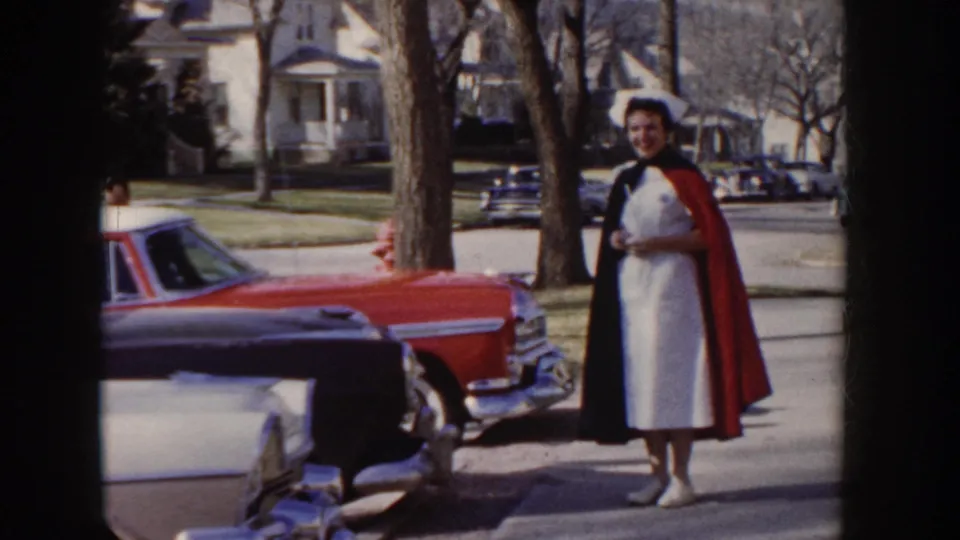 1955 Idaho: nursing graduate in white dress, dark cape, and nurse's cap stands beside a large-finned red American sedan on a tree-lined suburban sidewalk.