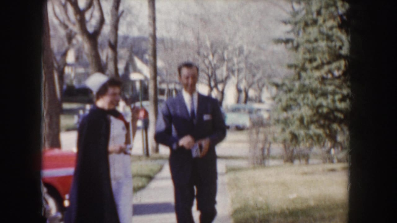 1955 Idaho: man in dark suit and nursing graduate in dark cape approach the camera operator on a tree-lined suburban sidewalk, red 1950s car visible at left.