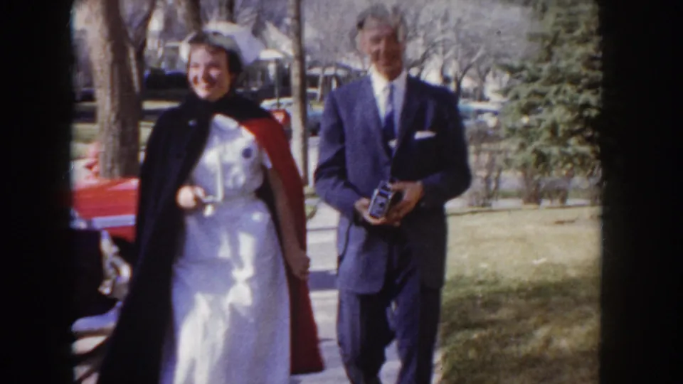 1955 Idaho: nursing graduate in white uniform and open dark cape walks with a suited man holding a box camera; a red 1950s sedan and suburban houses are visible in the background.