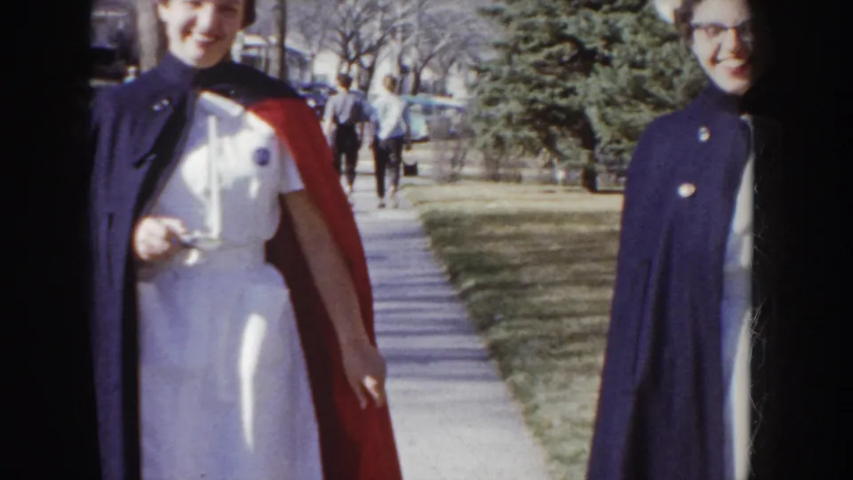 1955 Idaho: nursing graduate in dark navy cape with red lining, nurse's cap, and glasses smiles at the camera on a suburban sidewalk, second pedestrian visible behind.