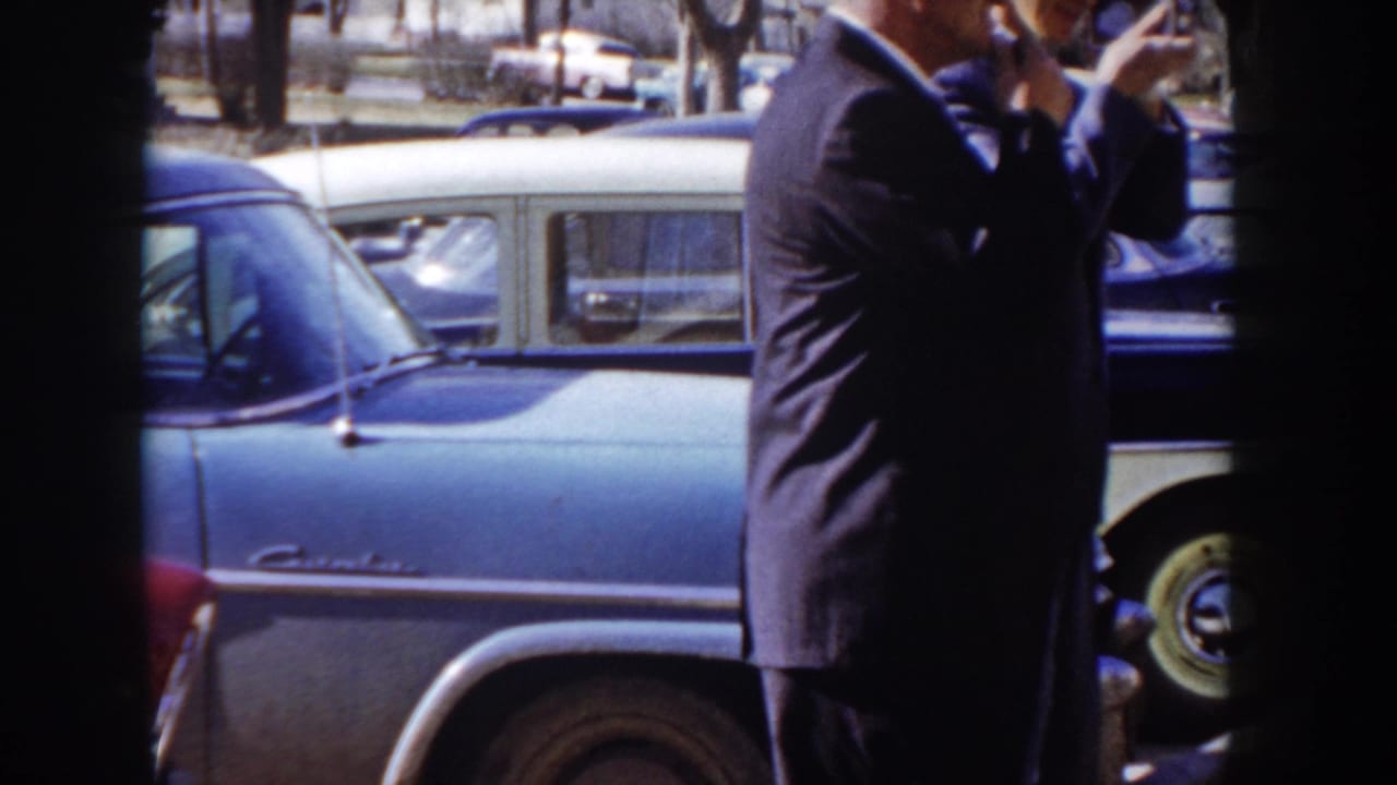 Rear view of a man in a dark suit holding a hand-held camera beside a large two-tone blue mid-1950s sedan; Idaho street, circa 1955 per metadata.