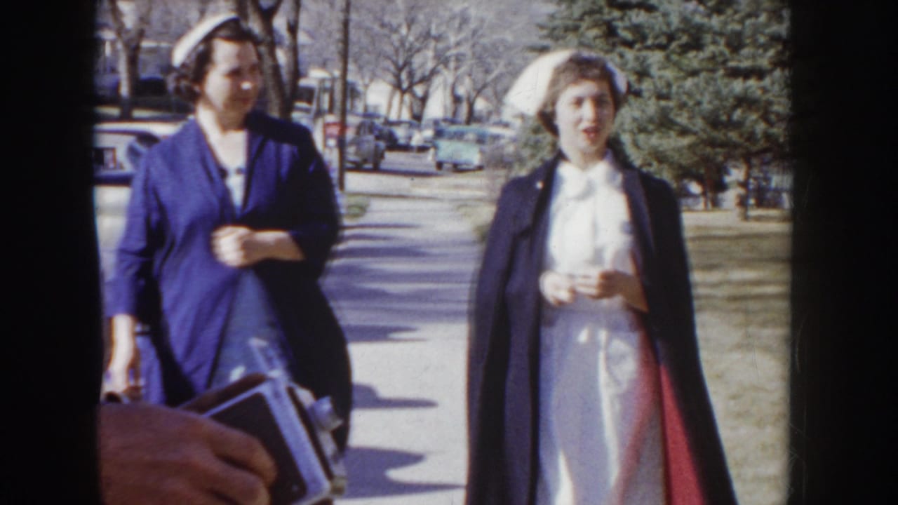 Two women in blue nursing uniforms and white caps walking on a sunlit sidewalk, one carrying a bag, with mid-1950s cars parked in background.