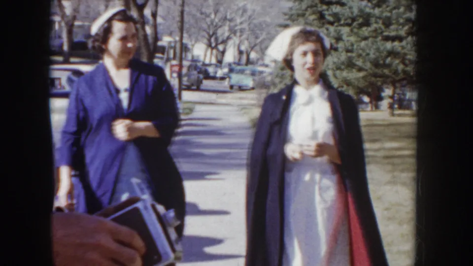 Two women in blue nursing uniforms and white nursing caps on a sunlit sidewalk, one carrying a dark bag; bare trees and period cars visible behind; Idaho, circa 1955 per metadata.