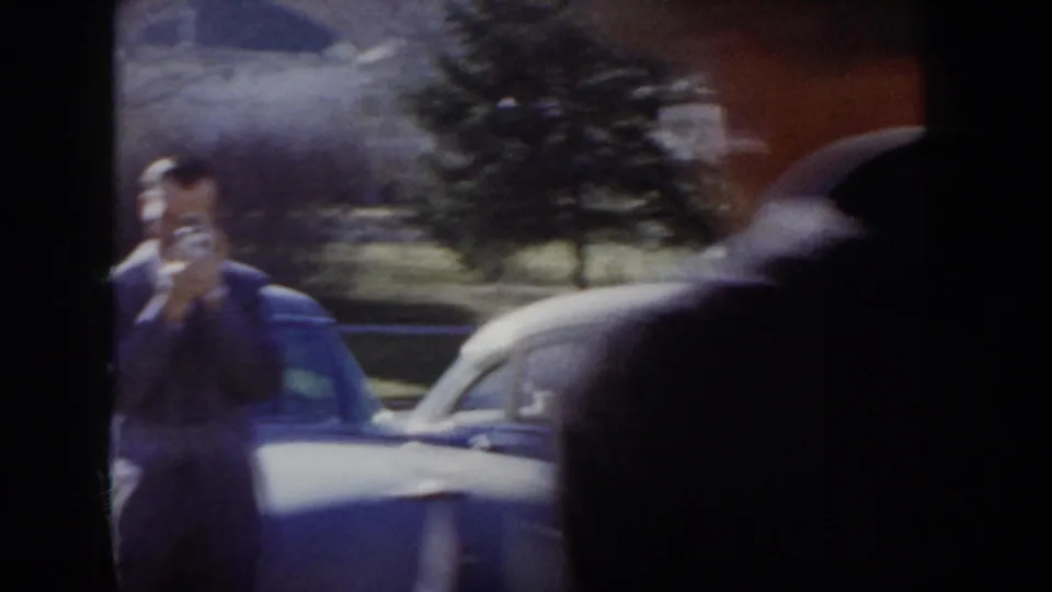 Man in a dark suit raising a hand-held film camera to his eye beside a mid-1950s sedan; evergreen trees behind; Idaho street, circa 1955 per metadata.