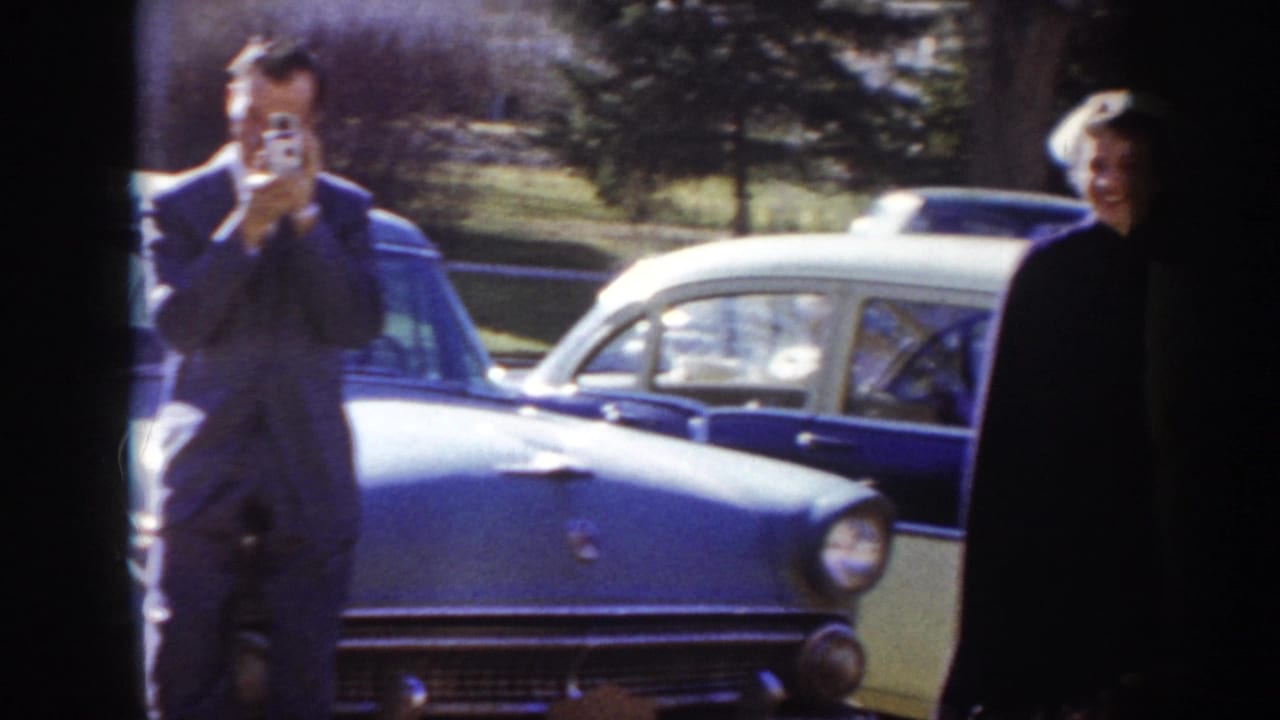 A man raises a small handheld camera to film beside a blue classic American car, with a second figure in lighter clothing partially visible at right, circa 1955 Idaho.