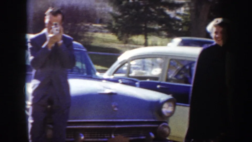 A man raises a small handheld camera to film beside a blue classic American car, with a second figure in lighter clothing partially visible at right, circa 1955 Idaho.