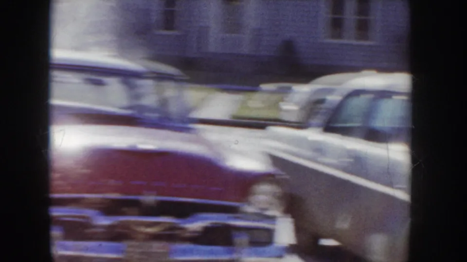 Motion-blurred pan across classic 1950s American automobiles on a residential street, red car visible at left, circa 1955 Idaho.
