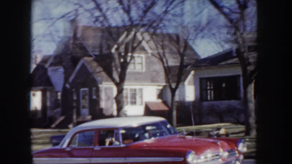 A red mid-1950s American automobile on a quiet residential street lined with two-story clapboard houses and bare trees, Idaho, circa 1955.