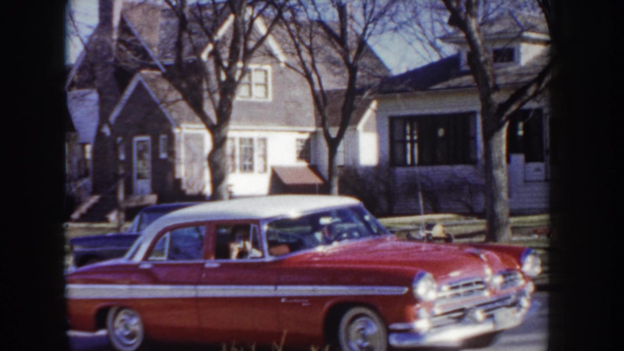 Mid-1950s two-tone red and white sedan with chrome grille and whitewall tires approaching camera on a tree-lined Idaho residential street in late autumn, c.1955.