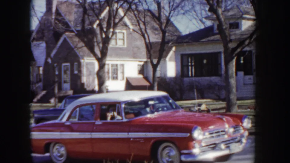 Mid-1950s two-tone red and white sedan with chrome grille and whitewall tires approaching camera on a tree-lined Idaho residential street in late autumn, c.1955.