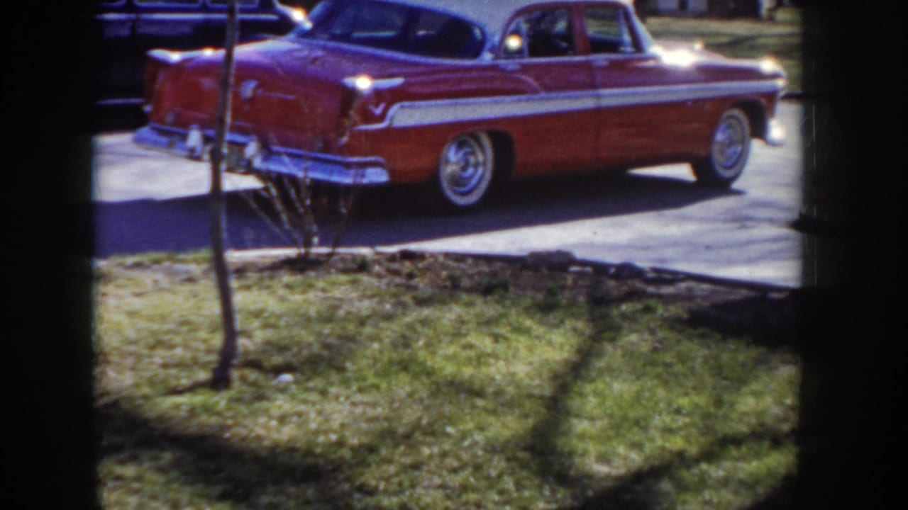 Close pass of a red and white 1950s American sedan, chrome trim and white roof stripe prominent, moving past the stationary camera.