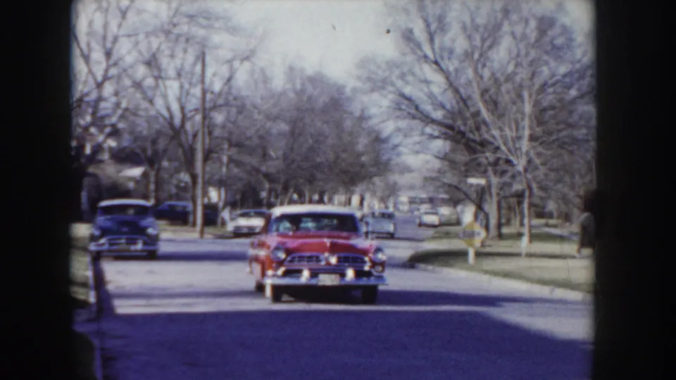 Mid-1950s two-tone sedan moving away from the camera down a tree-lined residential street, other period vehicles along the curb, bare autumn trees framing the scene.