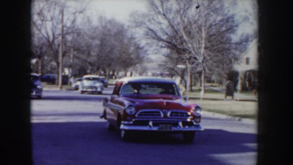 Mid-1950s two-tone red and white sedan departing down a tree-lined suburban street described as Idaho; bare branches and parked period cars line both sides of the road.