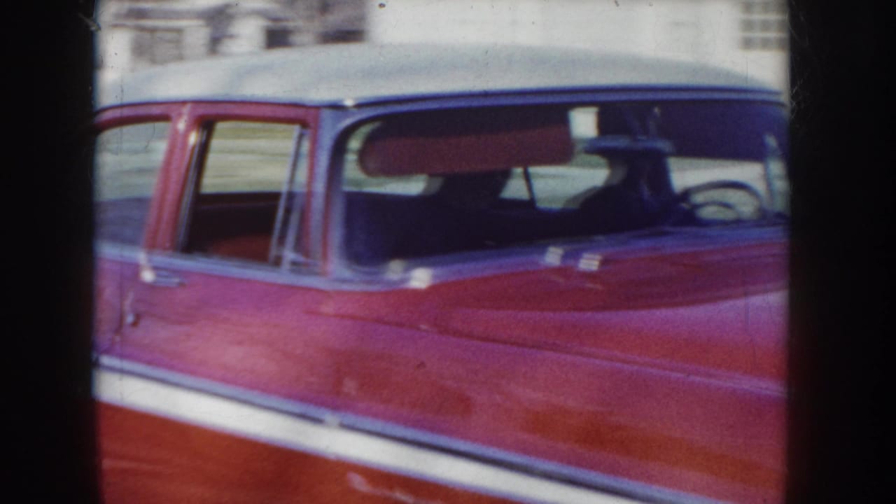 Close-up of the roof and upper body of a red and white two-tone 1950s sedan, showing chrome trim strip and rear window with small sticker.
