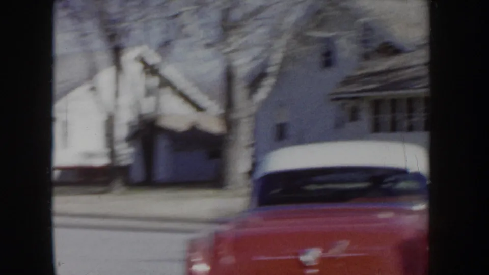 Red and white two-tone sedan passing modest residential homes and bare trees on a suburban street in Idaho, circa 1955; motion blur from camera pan.