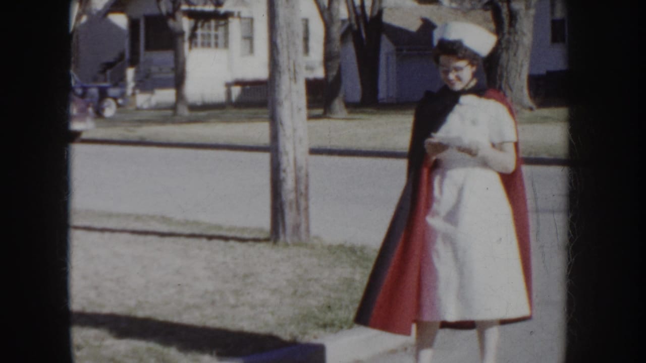 Close-up of a woman in a 1950s nurse's uniform — white dress, red cape, white cap — standing on a residential sidewalk, Idaho USA, circa 1955.