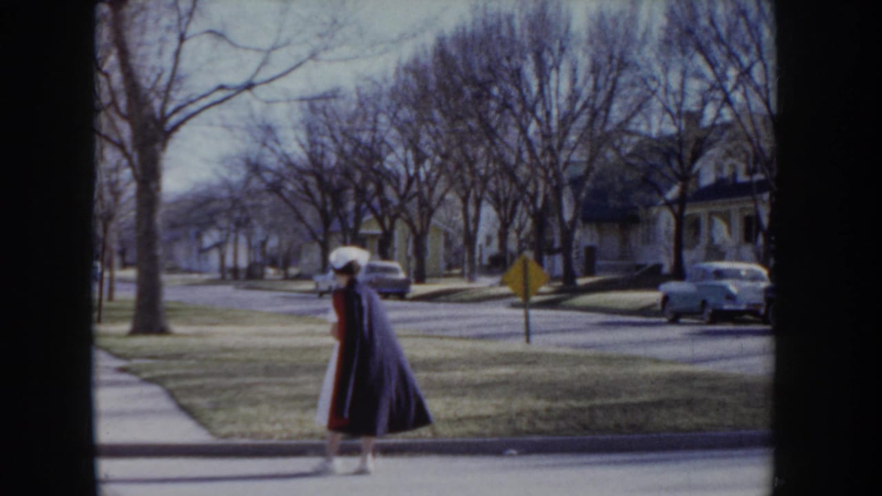 Nurse in white uniform and red cape walks along a tree-lined residential street; period automobiles and a yellow road sign are visible in the background.