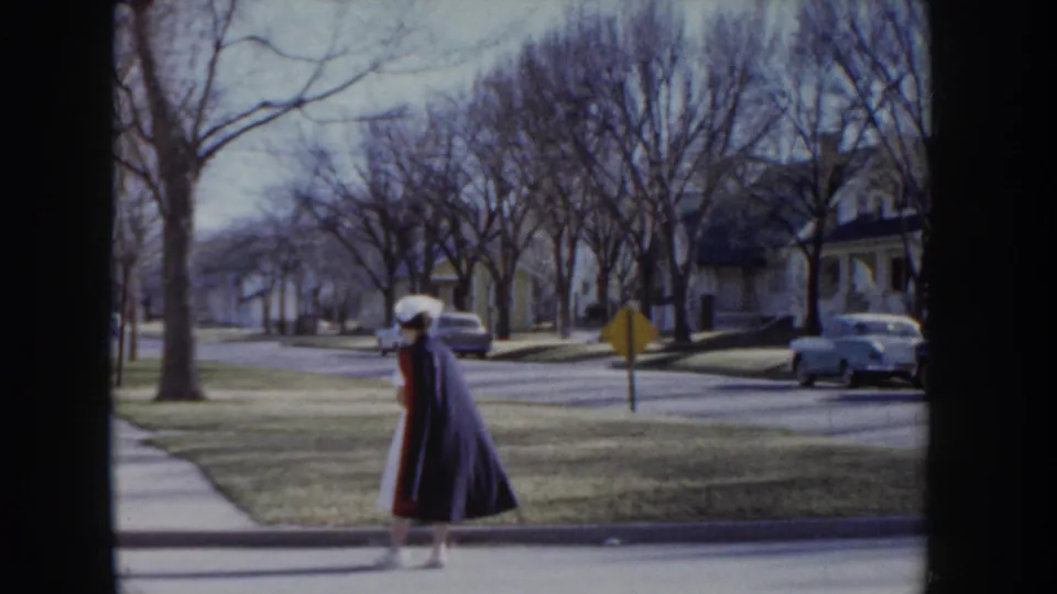 Wide shot of a nurse walking along a quiet tree-lined suburban street with 1950s-era parked cars and a yellow road sign visible, Idaho USA circa 1955.