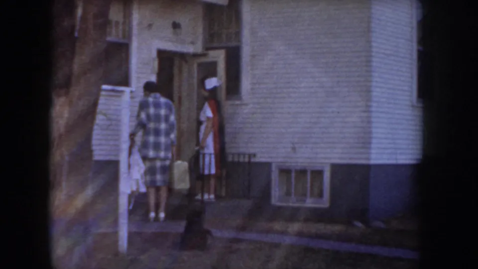 Nurse in white uniform and red cape with small bag is greeted by a man in a plaid shirt at the front door of a mid-century ranch home, Idaho circa 1955.
