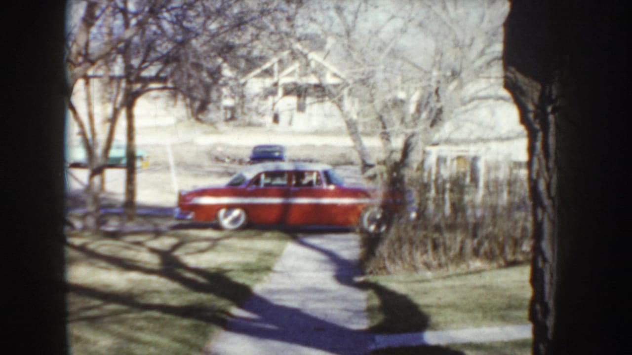 Wide establishing shot of a quiet Idaho suburban street circa 1955, with a red mid-century sedan, wooden fence, and long tree shadows on the pavement.