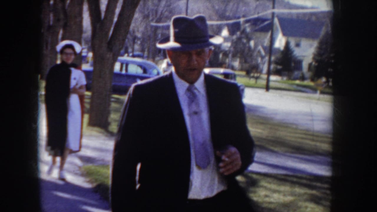 Man in dark suit and brimmed fedora walks toward camera on a suburban sidewalk; woman in white nurse's uniform stands in mid-ground behind him.