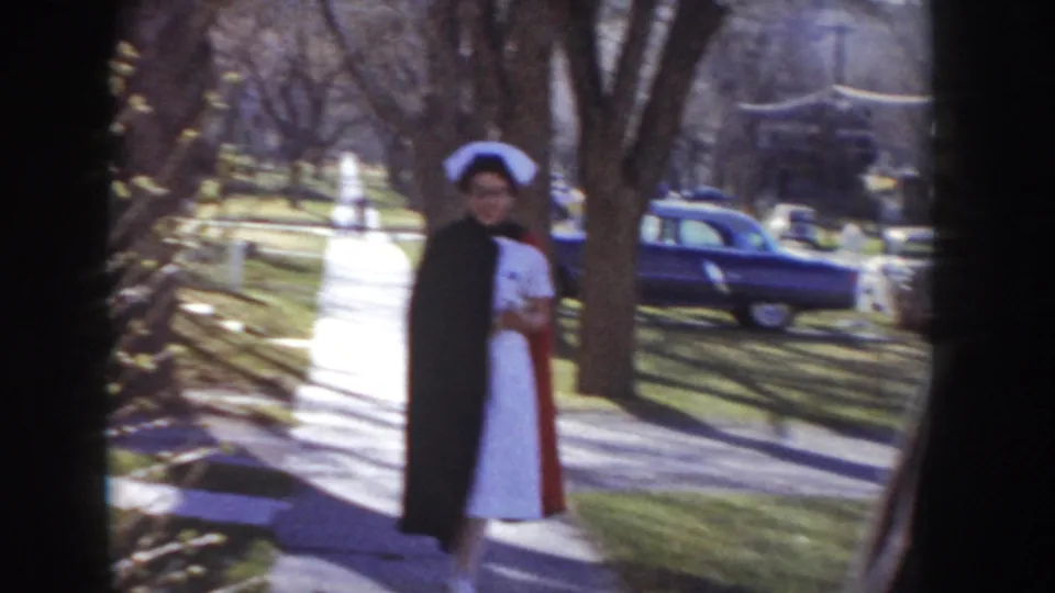 Woman wearing a white nurse's uniform, traditional cap, and dark outer coat stands on a residential sidewalk in 1950s Idaho, facing the camera.