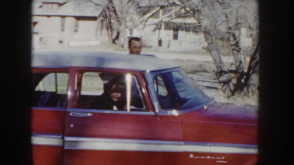 Close view of a red mid-century American sedan with chrome body trim on a 1950s Idaho residential street; a man in a hat stands at the door as a second figure appears through the window.