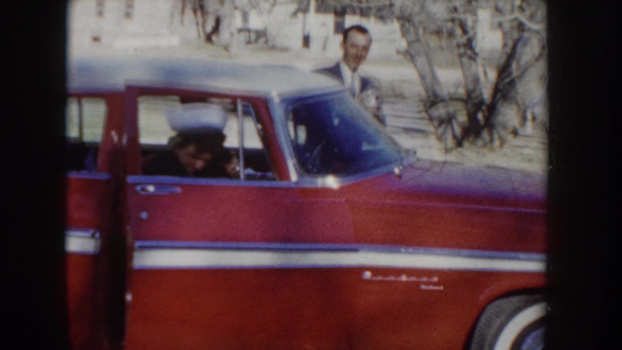 A man in a dark suit stands near a red two-tone American car in Idaho circa 1955, with a woman in a white nurse's cap visible inside or exiting the vehicle.