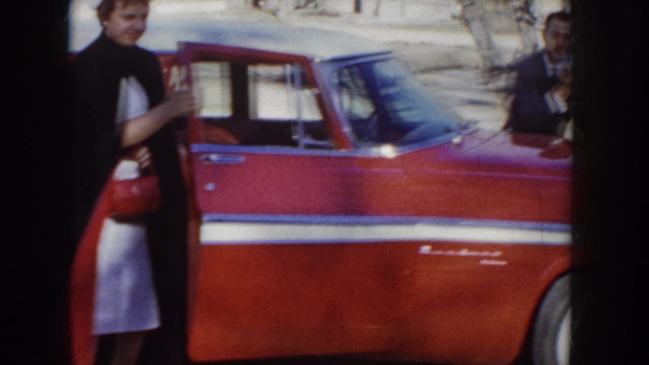 Woman in white nurse uniform and dark cape stands at the open rear door of a red American car; a second figure is visible at right edge.