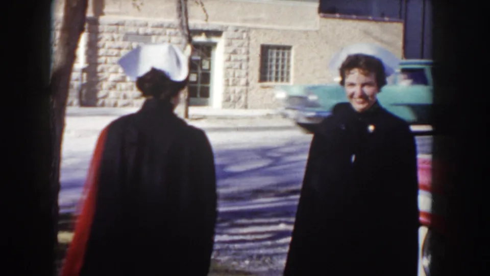 Two nurses in dark capes and white caps greet each other on a street in Idaho circa 1955; one smiles toward the camera with a stone building and a teal mid-1950s car visible behind them.