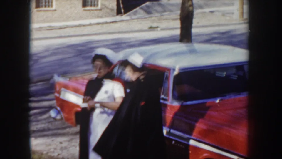 Two figures share a close greeting beside a red American sedan in Idaho circa 1955; one is in a white nurse's dress and cap, the other in a dark coat, with a stone building in the background.