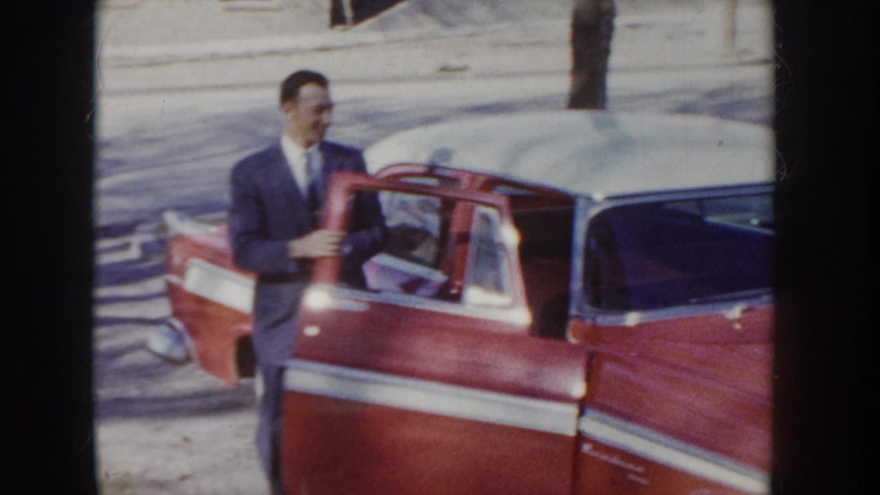 Man in a suit stands laughing beside the open door of a red two-tone 1950s sedan; a second figure is visible in the background.