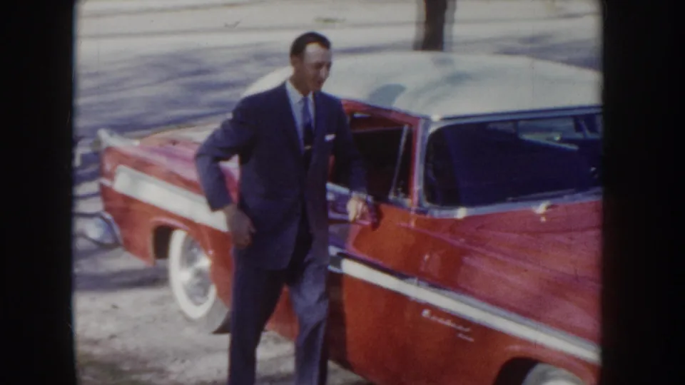 Man in dark suit poses by the open door of a red American sedan with prominent white-wall tires and chrome trim, circa mid-1950s Idaho.