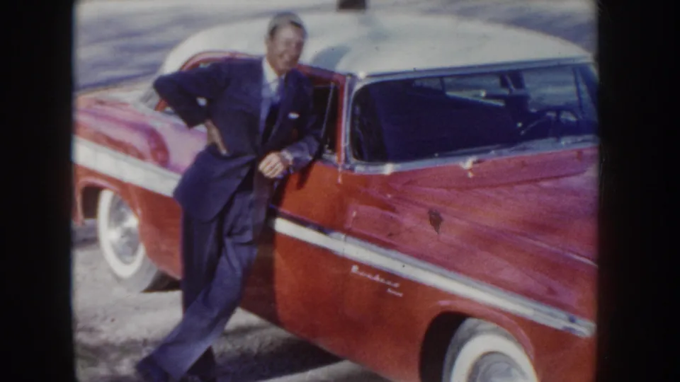 Man in dark suit leans casually against a red and white two-tone American sedan with chrome accents and white-wall tires, circa mid-1950s Idaho.