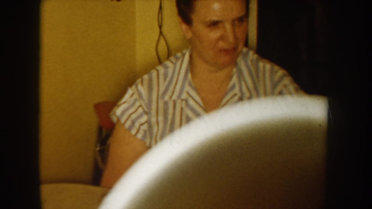 Opening frame of a mid-1950s 8mm home movie; woman in striped blouse visible behind a large white foreground object, likely a cup held close to camera; yellow wall behind.