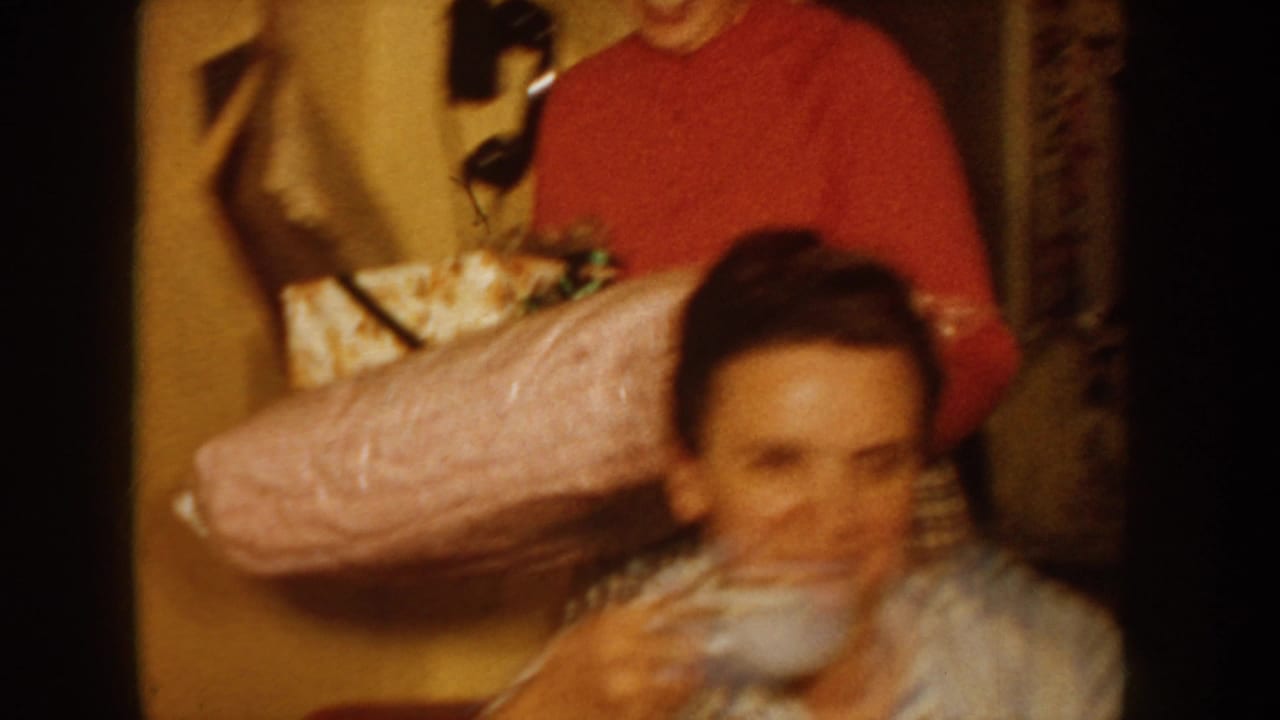 Two women in a home interior; one in a red sweater presents a large pink-wrapped gift to a seated woman with dark hair.