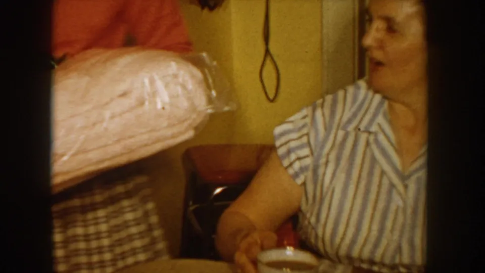 Woman in a blue-and-white striped blouse reacts with open mouth to a large pink-wrapped gift in what appears to be a 1950s home interior; telephone cord and plaid skirt of the gift-giver visible.