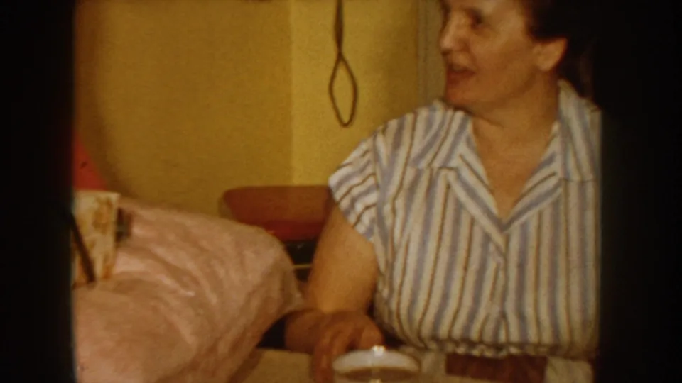 Woman in a blue-and-white striped blouse smiles warmly beside a large pink-wrapped gift in what appears to be a 1950s American home interior; yellow wall and telephone cord visible behind her.