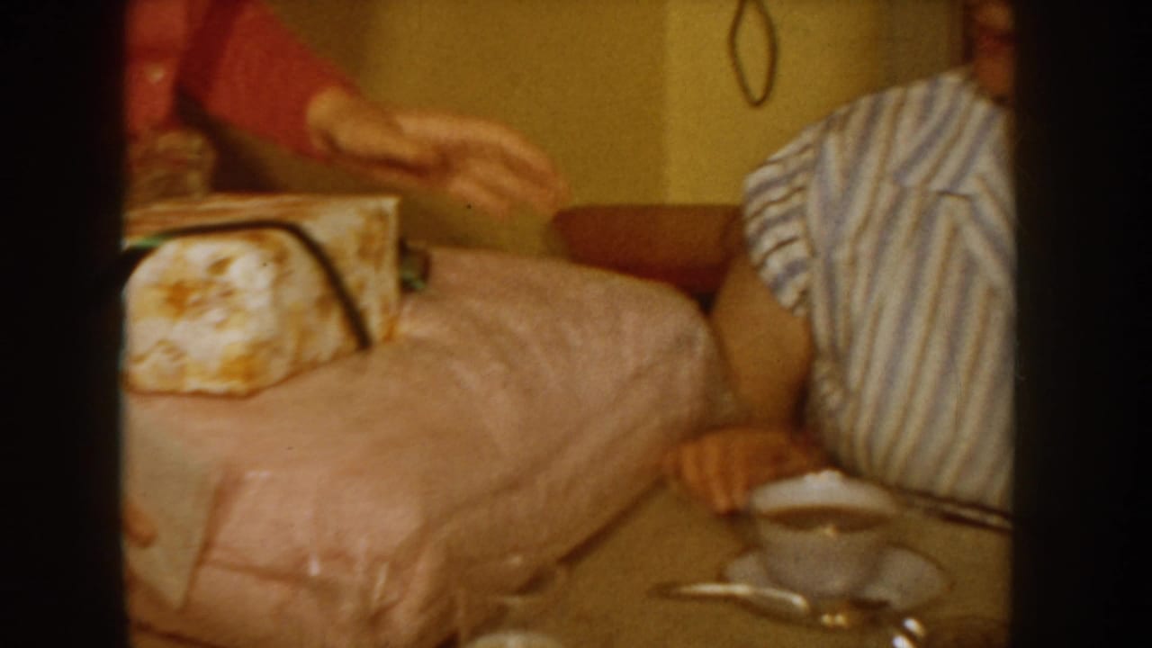 Two people in a domestic interior, one in blue-white striped blouse and one in red sweater, with a floral-wrapped gift on a pink bedspread, Idaho, USA, circa 1955.
