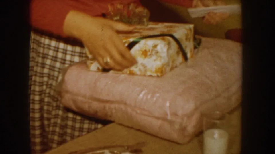 Close-up of hands unwrapping a floral-patterned gift box with a dark ribbon on a pink bedspread, circa 1955 Idaho domestic interior.