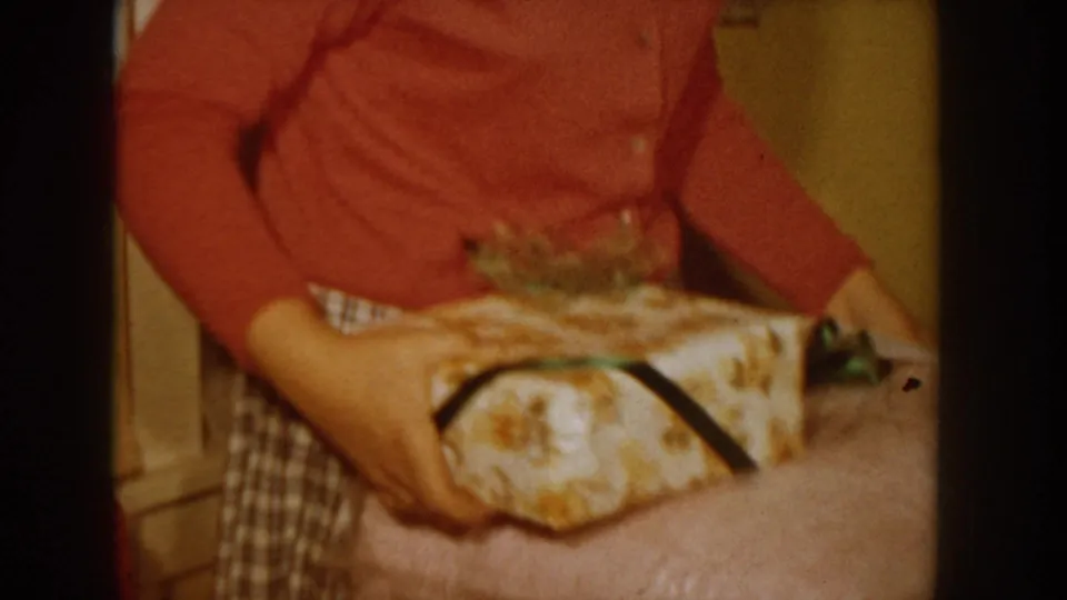 Person in red knit sweater and plaid skirt holds a floral-wrapped gift box with dark ribbon; a small green item is partially revealed, circa 1955 Idaho home interior.