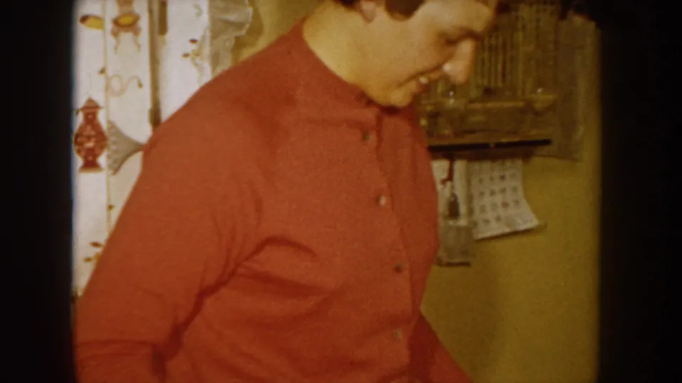 Close-up of a smiling woman in a red button-up sweater in a domestic interior, with floral curtain and wicker basket visible, Idaho, USA, circa 1955.