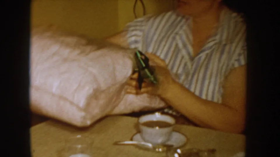 Tight shot of a woman in a striped blouse handling a large pink gift parcel; a white coffee cup and saucer are visible on the table surface, circa 1955