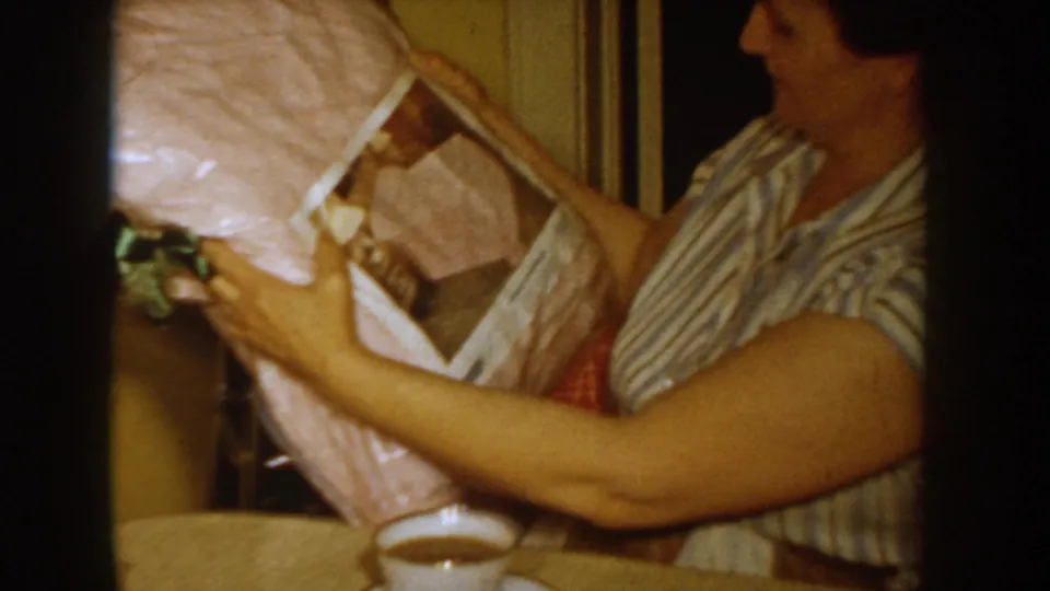 Woman pulls back pink wrapping paper to reveal a green bow or decoration and box contents beneath; a coffee cup is visible at the lower edge, circa 1955