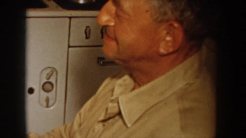 Close profile of a man in a light beige short-sleeve shirt watching off-screen; a vintage kitchen appliance with a period-style rounded latch is visible behind him, circa 1955