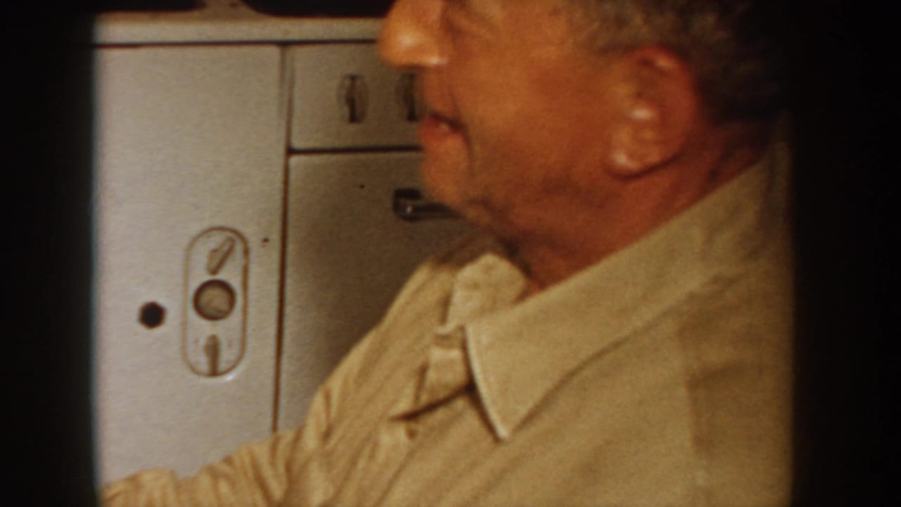 Close profile of an older man in a light tan collared shirt in a 1955 Idaho home; large white appliance with period controls visible at left.