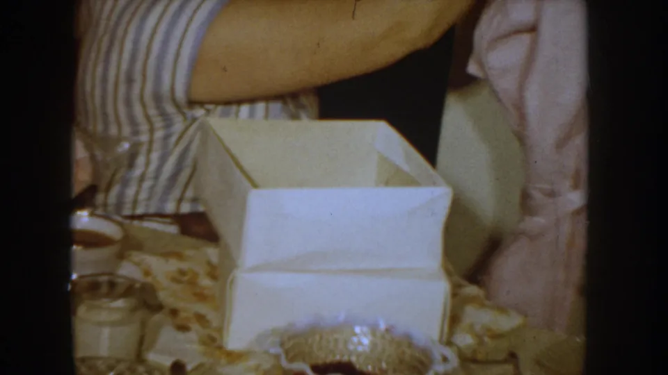 Open white gift box and tabletop items in a 1955 Idaho home interior; woman in blue-and-white striped dress at left edge of frame.