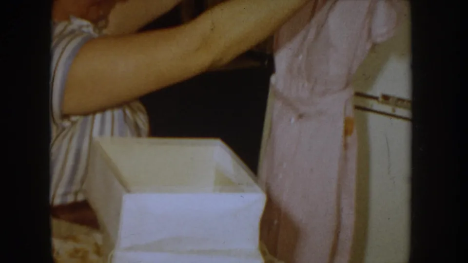 Woman in blue-striped dress with pink garment near open white gift box in a 1955 Idaho home interior; white mid-century appliance at right edge.