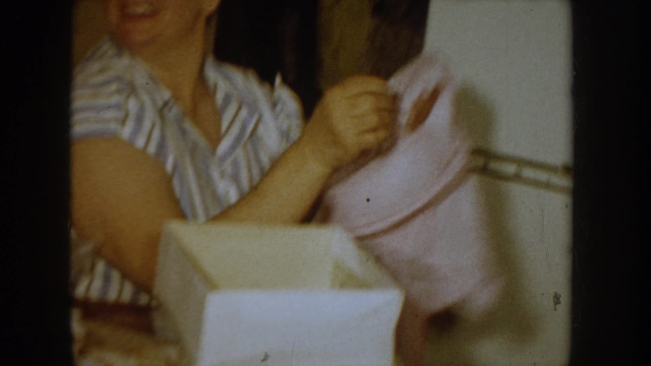 Close-up of a woman in a 1950s striped house dress lifting a pink clothing item from an open white gift box; Idaho, c.1955, 8mm.